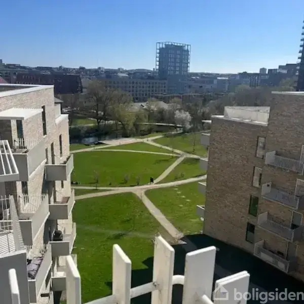Bygninger omgiver en park med stier, træer og græsplæne. I baggrunden ses flere højhuse og bylandskab med klare blå himmel. Pejsende sollys skaber skarpe skygger i området.