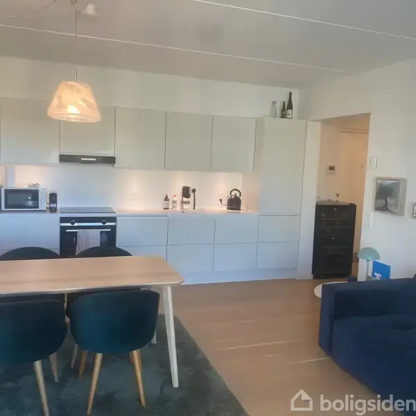 Et moderne køkkenalrum med hvidt køkkenmodul, mikroovn, indbygningsovn og kaffemaskine. Et spisebord med fire mørke stole står på et tæppe. Til højre er en mørk sofa.