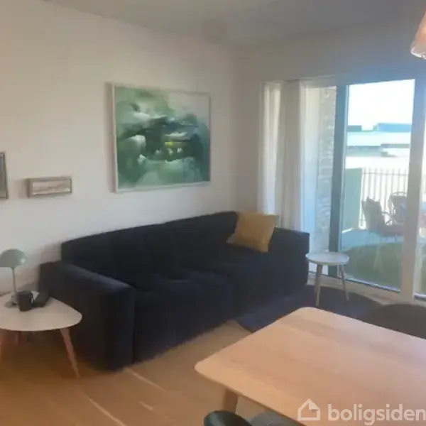En mørk sofa med en gul pude står på et trægulv ved siden af et rundt hvidt bord med en lampe. Store vinduer viser en balkon, hvor der står en stol. Malerier hænger på væggen.