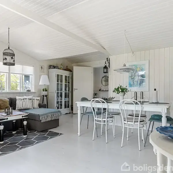 Stue med lyse møbler, herunder en sofa og spisebord med stole. Stuen har store vinduer, der giver naturligt lys, og udsigt til en grøn have. Væggene og loftet er hvide.