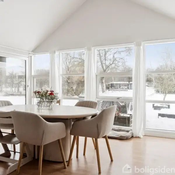 Et rundt spisebord med fem stole står i et lyst rum. Bordet er pyntet med en blomsterbuket, og store vinduer afslører en snedækket have udenfor.
