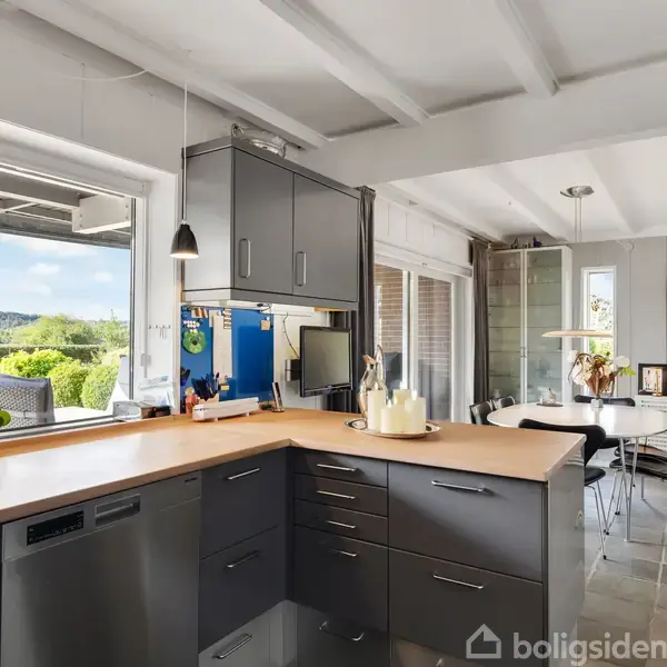 Et lyst, moderne køkken med grå skabe og en træbordplade. Det åbner ud mod en spisestue med et bord og stole. Store vinduer giver udsigt til en grønt udendørsområde.