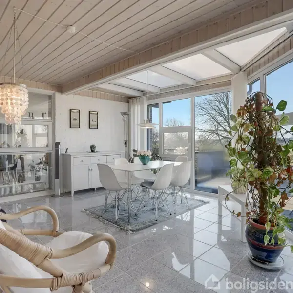 En lys, moderne stue med højt til loftet. En stor plante står ved gulv-til-loft-vinduer. Et spisebord med fire stole står nær en kommode og glasvitrineskab.