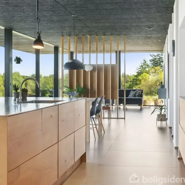 Køkken med træskabe og en sort bordplade, stående i forgrunden. Store vinduer slipper lyset ind og giver udsigt til grønne træer udenfor. Moderne indrettet.