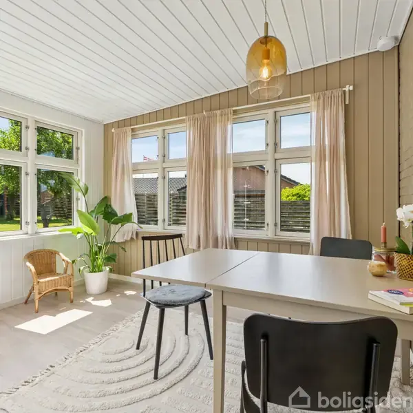En lys, indbydende stue med et bord og stole. Et vinduesparti slipper sollys ind. Grønne planter og en lænestol skaber en hyggelig atmosfære.