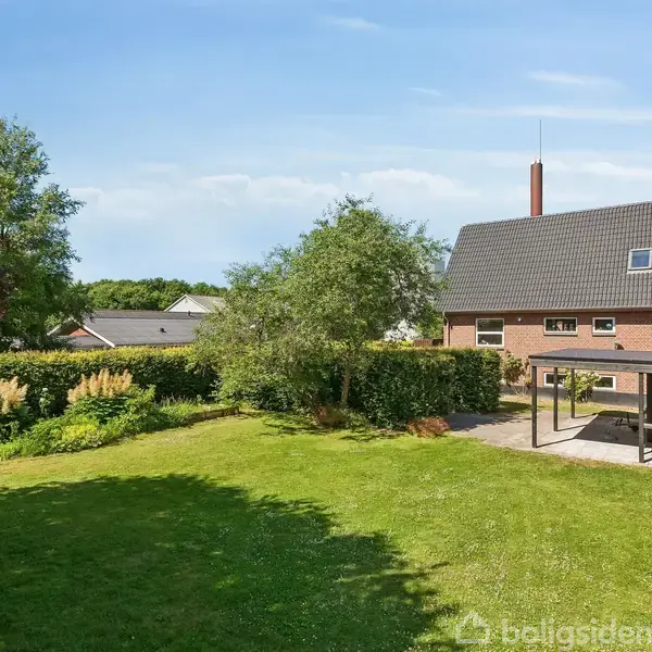 Huset er en murstensbygning med mørkt tag, omgivet af en stor have med græsplæne og træer. Til højre er en overdækket terrasse i en rolig villakvarter.