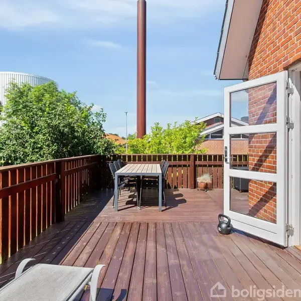 En træterrasse med bord og stole, omgivet af et hegn. Der er en åben dør fra et murstenshus, sollys skinner, og omgivelserne inkluderer grønne træer og en bygning i baggrunden.