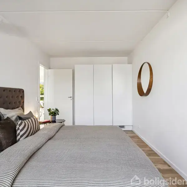 Soveværelse med en stor seng dækket af grå og stribede puder. En rund spejl hænger på væggen ved siden af en hvid garderobe. Dør står åben med udsigt til planter.