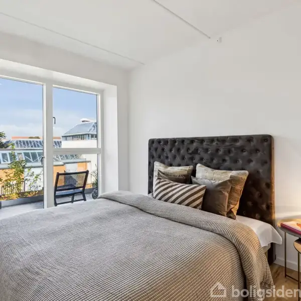 En seng med gråt sengetøj og puder står ved siden af et natbord med lampe og plante. Store vinduer åbner ud til en balkon med udsigt over nabotagene.
