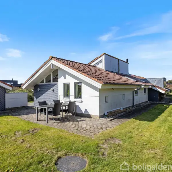 Hvidt hus med rødt tegltag, står på en solrig græsplæne med flisebelagt terrasse. To stole og et bord ses foran huset. Blå himmel og en garage i baggrunden.