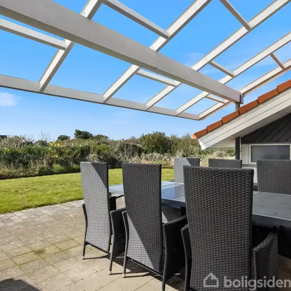 Udendørs spisebord med otte høje stole står på en terrasse under en pergola ved siden af et hus. Baggrunden viser en velplejet græsplæne og buskads under en klar blå himmel.