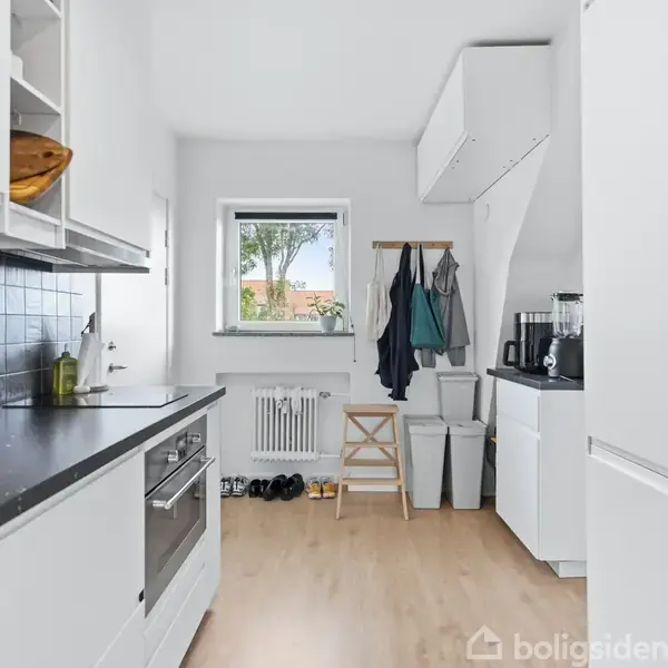 Et lyst køkken med hvide skabe og sorte bordplader. En ovn er indbygget, og der er en vask ved siden af. Vindue mod grønt område. Stumtjener og skoskab ved siden.