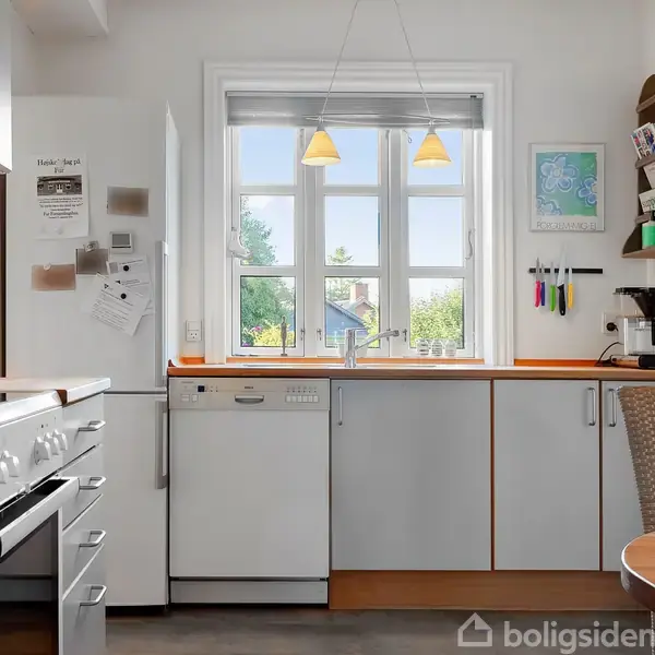 Et moderne køkken med lys grå skabsindretning og træborde. Opvaskemaskinen og komfuret er synlige. Et vindue med udsigt og lamper hænger over vasken. Noter og redskaber er på væggen.