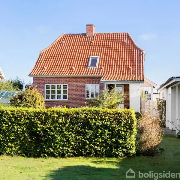 Rødt murstenshus med rødt tegltag står stille i en have med grønne buske og græsplæne, omgivet af enkelte nabo bygninger under en blå himmel.