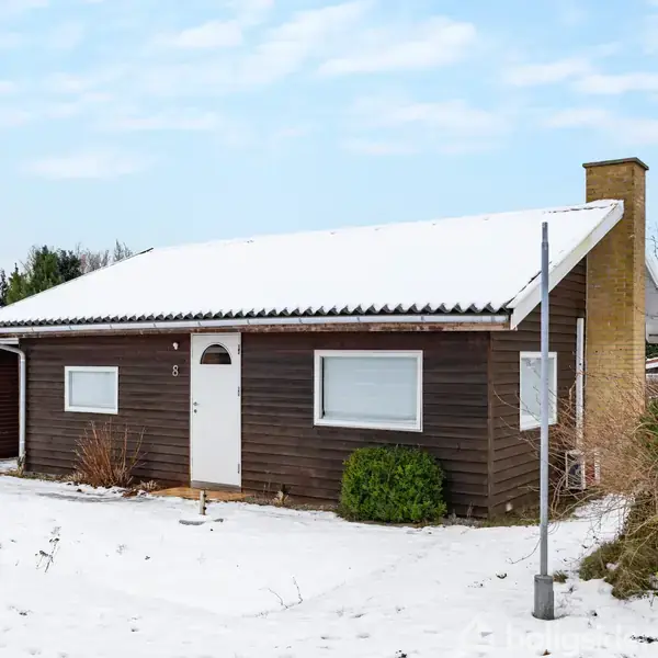 Et lille, brunt træhus står i sneen med en hvid dør og to vinduer. Omkring huset ses en snedækket grund og nogle buske. Nummer 5 er synligt på facaden.