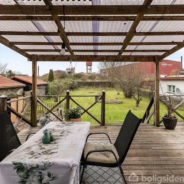 Terrassebord med dug og to stole. Bordet står under en træpergola. Baggrunden består af en have med græs, buske og et rødt bygningstag.