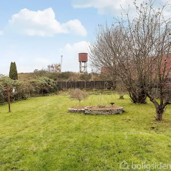 Grøn have med træer og buske står stille omgivet af et hegn; i baggrunden ses en rød bygning og en vandtank, mens himlen er blå med hvide skyer.