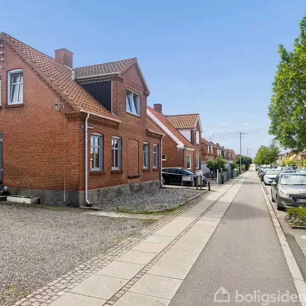 Rødt murstenshus med skråtag, står ved en stille vej med parkerede biler. Cykler står ved siden af huset. Fortov og træer danner rammerne på den anden side af vejen.