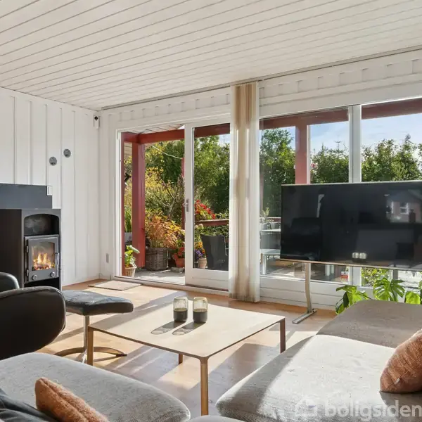 En hyggelig stue med en grå sofa, sofabord og stor fladskærms-TV. En pejs er tændt til venstre. Store vinduer bagved viser en grøn have udenfor.