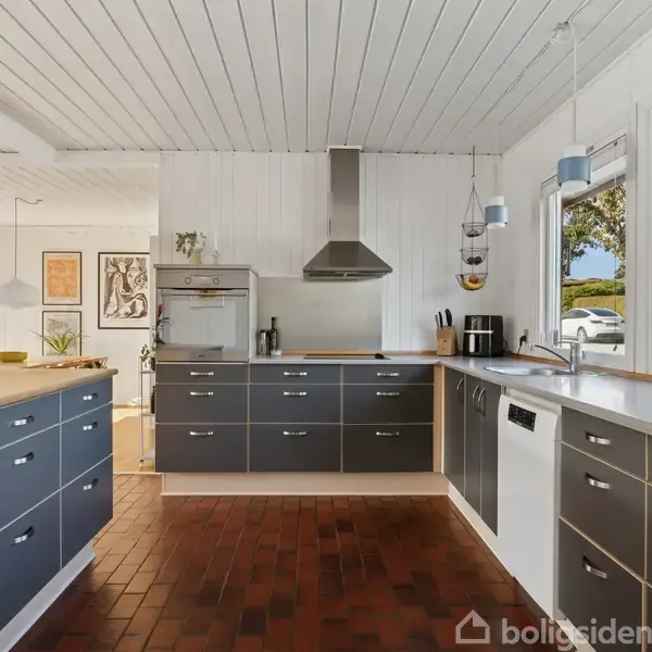 Køkkenet har grå skabe og træborde, med hvide vægge og loft. Køkkenudstyr inkluderer emhætte, ovn og opvaskemaskine. Rummet er lyst med store vinduer, planter, og hængende lamper.