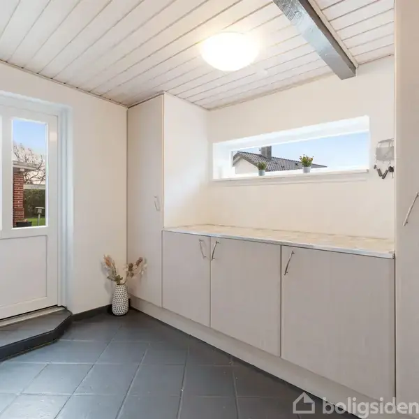 Et lyst rum med hvide skabe langs væggen, en rektangulær vinduesåbning ovenfor. En dør med kattelem fører udenfor. Rummet har mørke fliser og dekorative planter.
