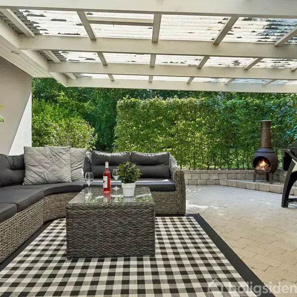 Udemøbler er arrangeret i et overdækket terrasseområde med en L-formet sofa og et glasbord. Et tæppe ligger under møblerne. En grill og udendørspejs ses mod en baggrund af grønne buske.