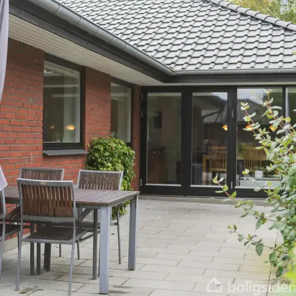Rundt havebord med stole står på fliser ved en murstensbygning. En parasol er opstillet ved siden af, mens planter omkranser området.