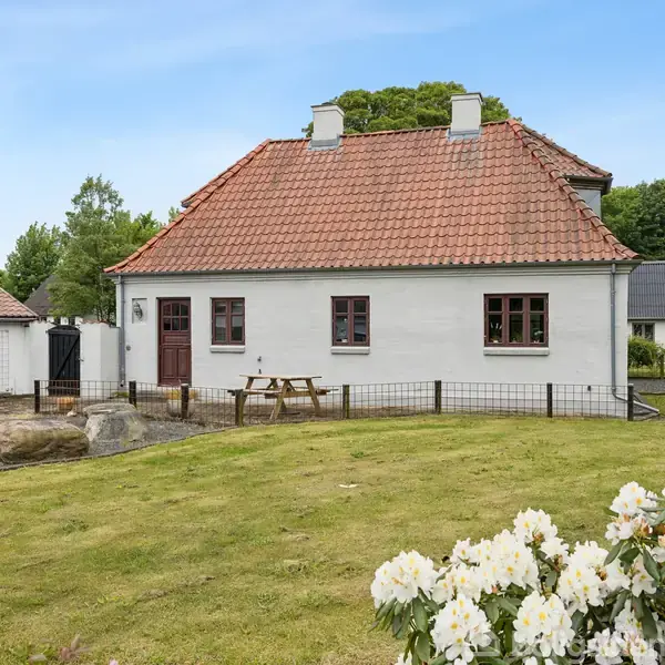 Hvidt hus med rødt tegltag står omgivet af en grøn plæne og buske. En brun dør og små vinduer ses på facaden. Træbord med bænke foran.