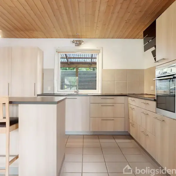 Lyst køkken med træloft, lyse træskabe og lyse fliser på gulvet. Elektrisk ovn indbygget i skab. Høj stol ved køkkenbordet. Vinduet viser en træterrasse udenfor.