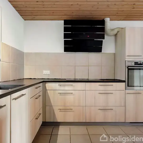 Køkken med lyse trælignende skabslåger og sorte bordplader står stille i moderne stil. Vask og vindue til venstre, ovn og emhætte. Loftet er træbeklædt.