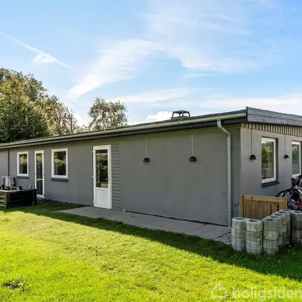 En grå bungalow står stille i et velholdt haveområde med græsplæne og en grusbelagt indkørsel. Cykler er placeret ved siden af huset.