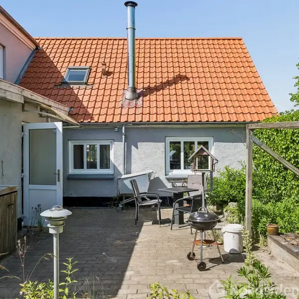 Baggård med flisebelægning viser havemøbler og en grill. Bygningen har rødt tegltag og grå vægge. Omkranset af grønne buske og planter. Solrigt vejr skaber en lys stemning.
