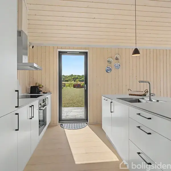 Et moderne køkken med hvide skabe og køkkenø. Sollys strømmer ind gennem en glasdør, der fører ud til en græsplæne. Træbeklædning på væggene.