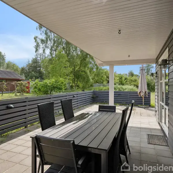 Langt, sort havebord står på en terrasse med fliser. Seks stole omgiver bordet. Kontekst er en lukket terrasse med træhegn, omgivet af træer og en blå himmel.