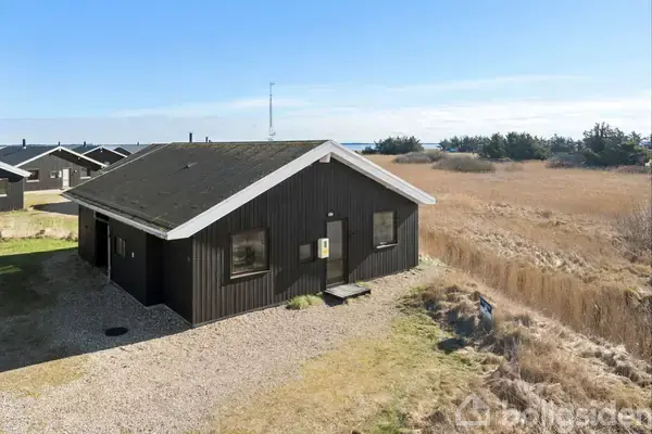 Sort træhus står stille på en grusbelagt grund omgivet af tørt græslandskab og flere lignende huse i baggrunden under en klar blå himmel.