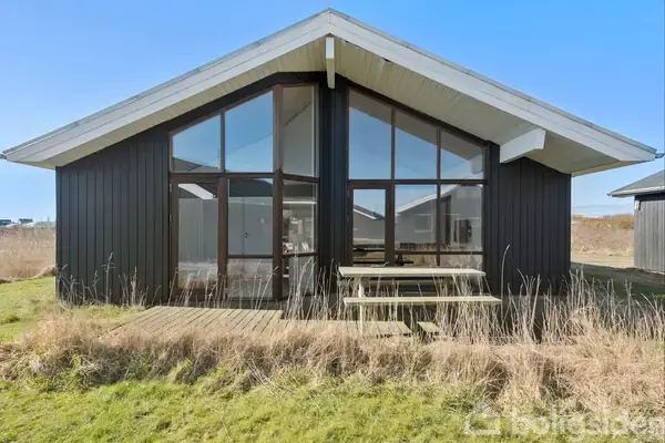 Sort træhus med store glasfronter står stille i en vildtvoksende have. Foran huset er en træterrasse med en bænk, og græsset er ujævnt. Himlen er klar og blå.
