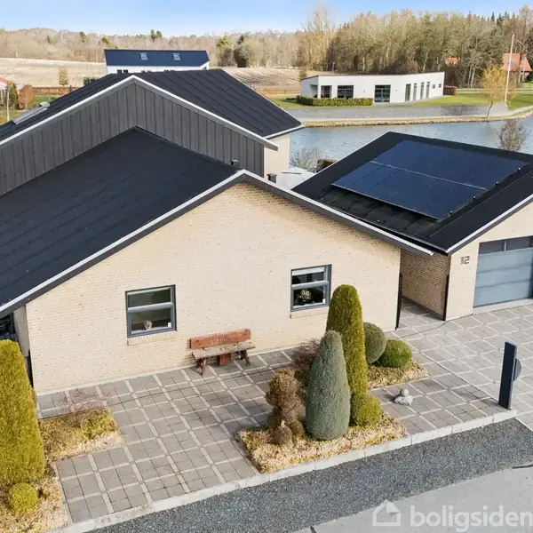 En moderne villa med solpaneler på taget står stille ved en flisebelagt indkørsel. Omkring huset er der trimmede buske og en baggrund med en sø og skov.