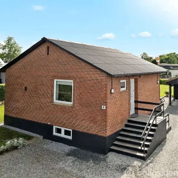 En lille, rød murstensbolig med en sort trappe, der fører op til hoveddøren, er omgivet af en grusindkørsel og grønne omgivelser med en garage i baggrunden.