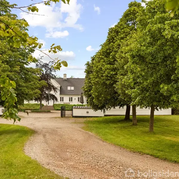 Et stort hvidt hus står for enden af en grusvej omkranset af grønne træer og velplejede græsplæner under en blå himmel med skyer.