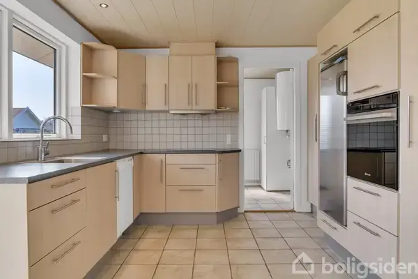 Køkken med beige skabe og hvide fliser; moderne apparater, indbygget ovn, opvaskemaskine og stort vindue; flisegulv og lys træloft i et lyst rum.