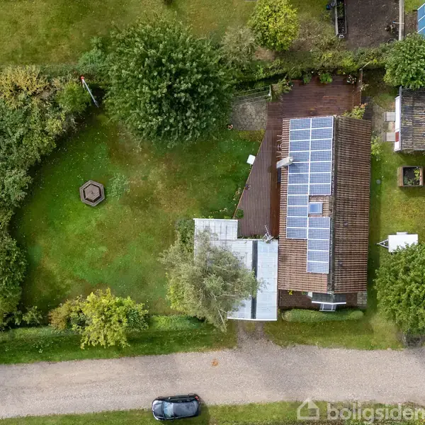 Et hus med solpaneler på taget står i en grøn have omgivet af buske og træer. Ved siden af huset er en lille bygning, og en bil er parkeret på vejen udenfor.