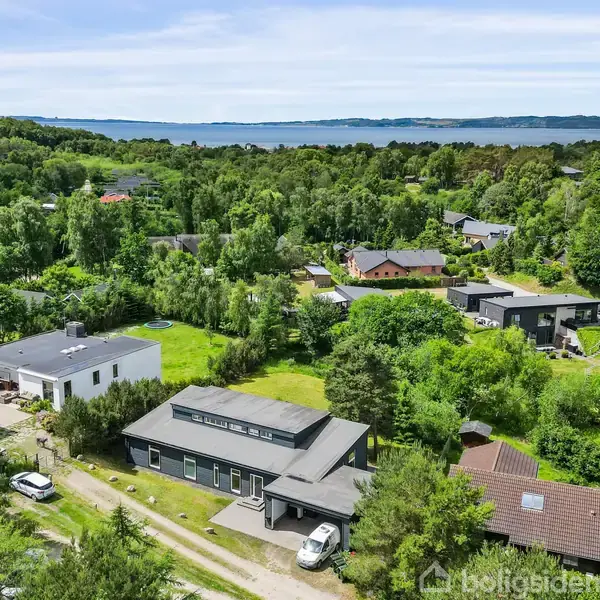 Hus omgivet af grønne træer, placeret tæt på en vej, med flere omkringliggende boliger. I baggrunden ses en udsigt over kyst og hav.