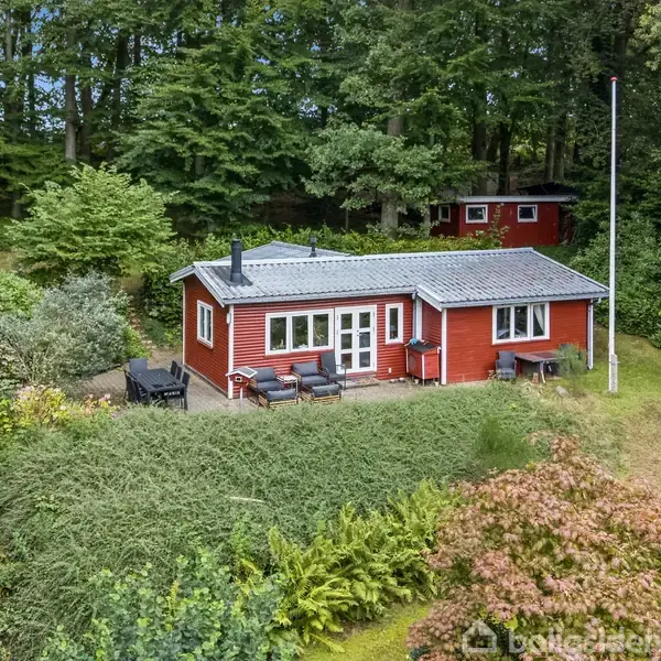 Et rødt sommerhus står i en frodig skovklædt have. Huset har en terrassedør med stole udenfor. Grønne buske og træer omkranser området, der er fredfyldt og naturligt.