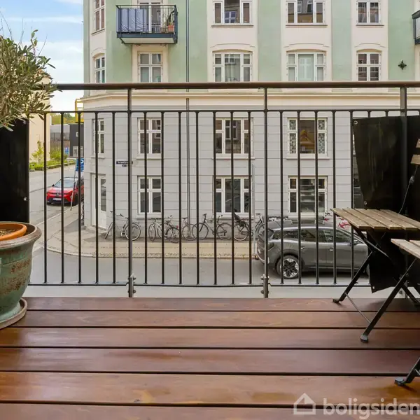 Balkon med træplanker, havemøbler og en stor plante i en krukke. Udsigt over gade med parkerede biler og klassiske lejlighedskomplekser i baggrunden.