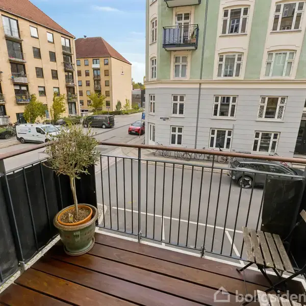 En balkon med trædæk, hvor en krukke med en plante og en lille stol står. Udsigt over en byvej med parkerede biler og lejlighedsbygninger i baggrunden.