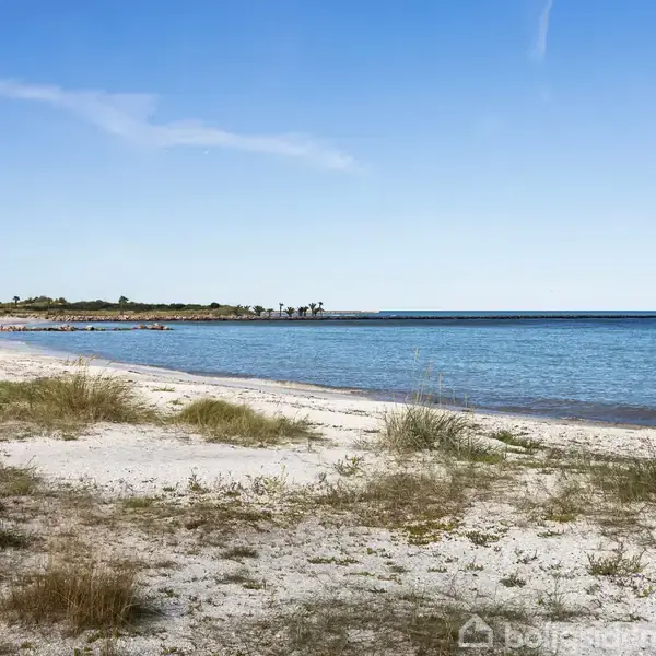 En øde strand med sand og spredt græs strækker sig langs kystlinjen. Roligt blåt hav møder horisonten under en klar himmel.