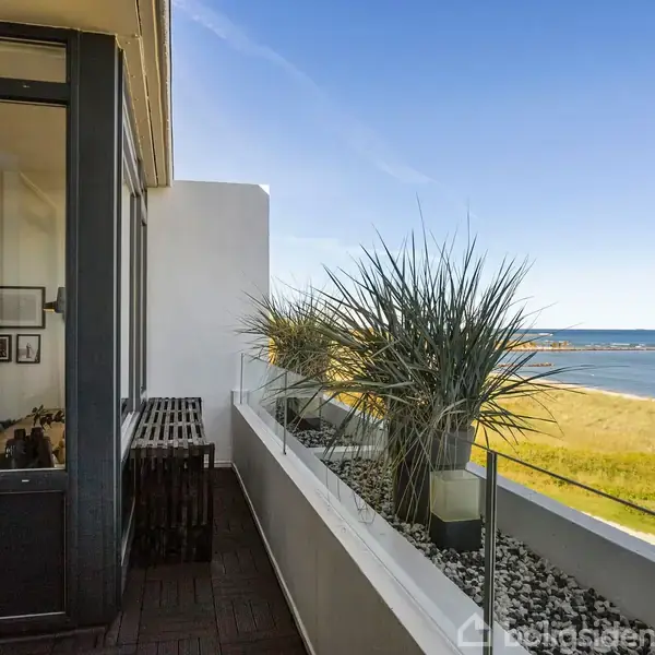 En altan med glasværn har planter i krukker; den leder til en stue gennem en dør. I baggrunden ses en strand med udsigt til havet under en klar blå himmel.