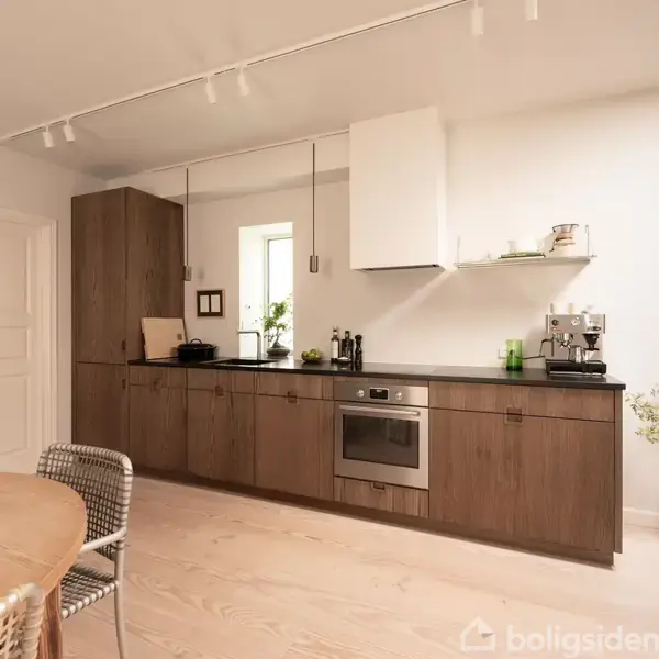 Et køkken skab i træ med en metalplade mærket "SIMONSEN CZECHURA COPENHAGEN". Skabet står i et lyst rum med en plante i vindueskarmen og et elegant, minimalistisk design.