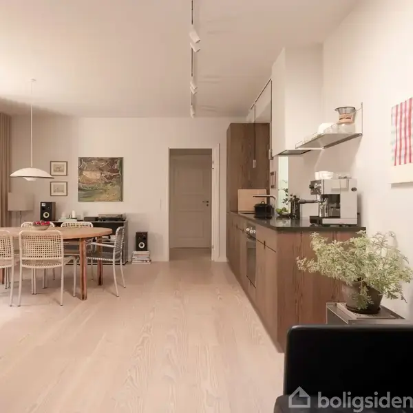 Sofabord med grøn plante står ved siden af køkkenområde. Spisebord med stole er bagved. Lyse trægulve og vægkunst supplerer det moderne interiør. Ingen tekst til stede.