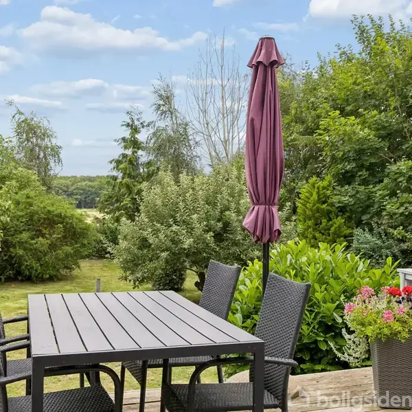 Et bord og fire sorte stole står på en terrasse med en sammenfoldet parasol. Omgivet af grønne buske og træer i en frodig have.